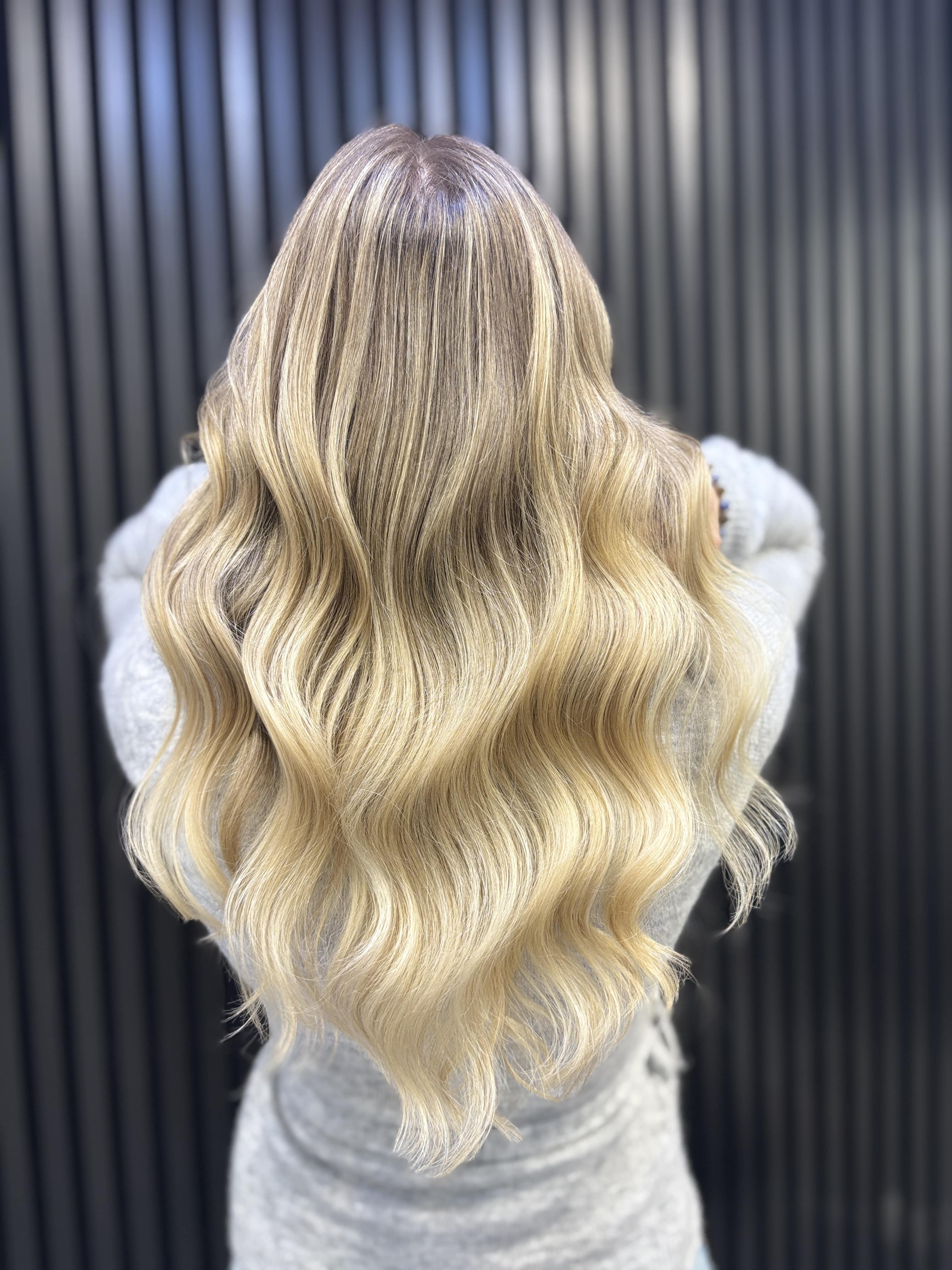 Eine Frau mit langen, gewellten, blond balayagierten Haaren steht vor schwarzer Wand